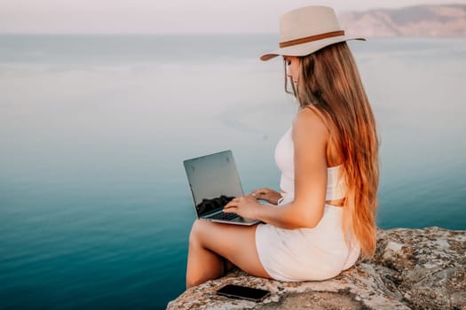 Digital nomad, Business woman working on laptop by the sea. Pretty lady typing on computer by the sea at sunset, makes a business transaction online from a distance. Freelance, remote work on vacation