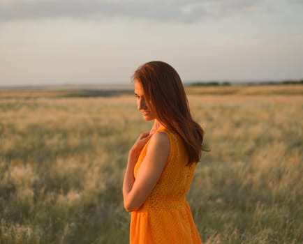 A pretty young girl with long brown hair in an orange dress in a field at sunset. The concept of freedom and nature