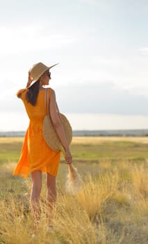 Beauty romantic girl outdoors. Rear view of a beautiful girl dressed in a casual orange dress with a straw hat and a straw bag in her hands in a field in the sunlight. Blows long hair. Autumn. Shine the sun, sunshine. backlit Warm tinted.