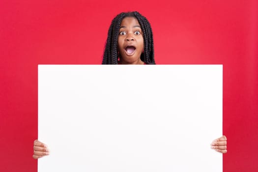 Surprised african woman holding a blank panel in studio with red background