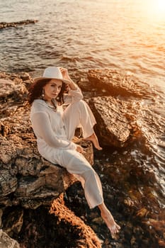 woman sea sunset. woman in a white pantsuit and hat is sitiing on the beach enjoying the sea. Happy summer holiday.