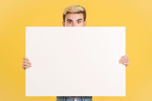 Surprised gay man hiding behind a blank board in studio with yellow background