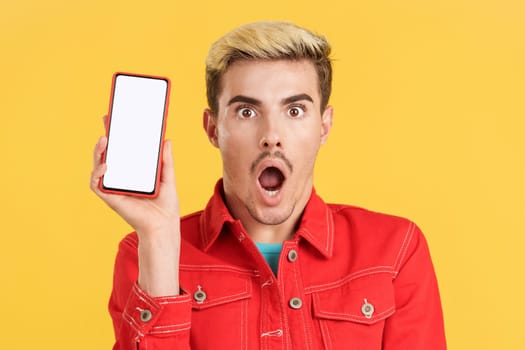 Surprised gay man holding a mobile with a blank screen in studio with yellow background