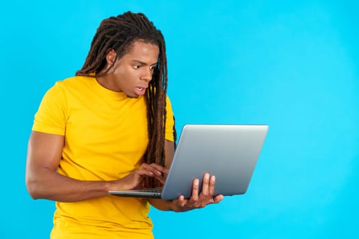 Surprised latin man with dreadlocks using a laptop in studio with blue background