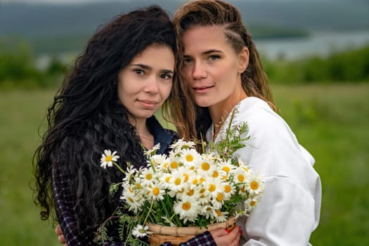 Two women enjoy nature in a field of daisies. Girlfriends hugging hold a bouquet of daisies and look at the camera