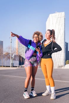 vertical photo of two female runners pointing ahead standing in the city ready to run, concept of friendship and sportive lifestyle, copy space for text