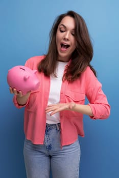 positive brunette young woman in a shirt and jeans puts her budget into a piggy bank.