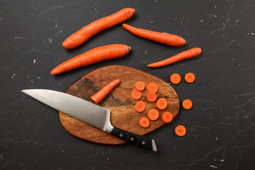 Top down view, chopping board, chef knife, and carrots, some of them cut in small circles, on black marble desk.