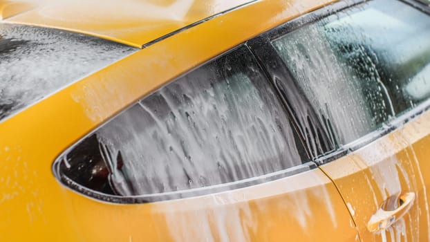 Yellow car washed in self serve carwash, shampoo spraying from hose to roof, streams of foam flowing down the window.