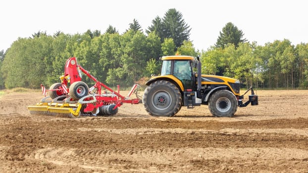 Yellow tractor pulls red sowing mechanism over dry field, trees in background.