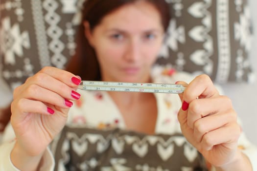 Young sick woman laying in bed, holding classic mercury thermometer in front of her.