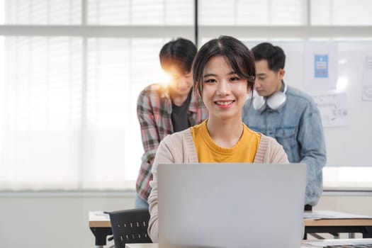 young Asian female marketing assistant is working on her work on her laptop at her desk while her coworkers are working in the room