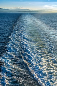 Foamy trail on water surface behind a big ocean vessel. Coastal Mountains view from a ship on Pacific ocean.