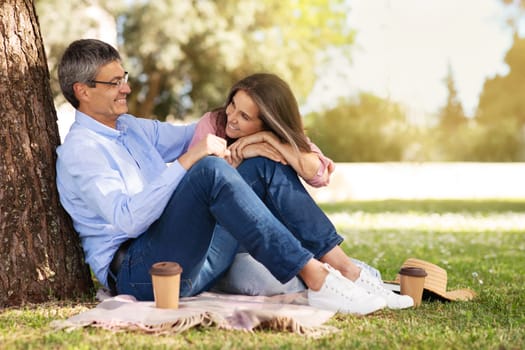 Loving Mature Couple Relaxing Together Outdoors In Summer Park, Happy Romantic Middle Aged Spouses Sitting On Plaid Under Tree And Drinking Takeaway Coffee, Enjoying Picnic Outside, Copy Space