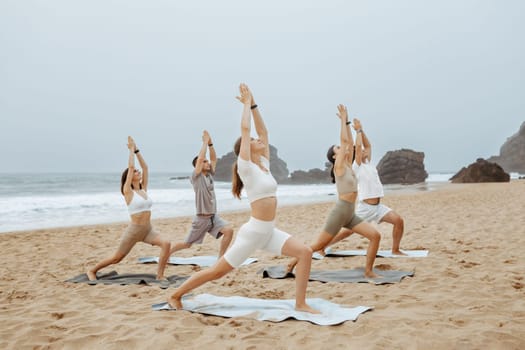 Yoga class on ocean shore. Group of young people doing warrior pose, meditating and exercising on the beach. Wellness and healthy balance lifestyle