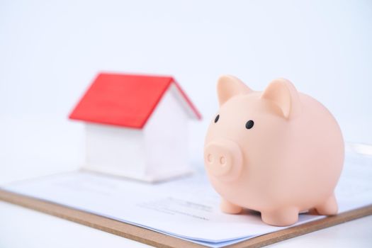 Piggy bank, beautiful wooden house model over a signed contract on white background, concept of saving money to buy a home insurance, close up, copy space.