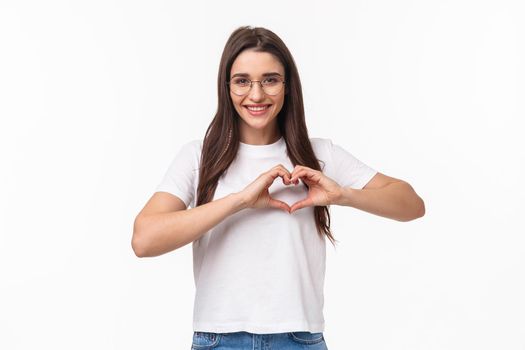 Portrait of caring and romantic lovely young woman in glasses, t-shirt, showing heart sign and smiling, express love care and sympathy, standing white background, open her heart to you.