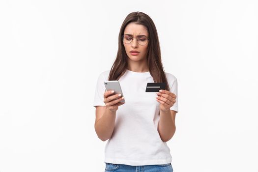 Portrait of serious-looking, focused young attractive woman copying digit numbers from credit card into billing info in app, paying or products online, purchase in internet store with smartphone.