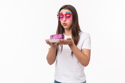 Entertainment, fun and holidays concept. Portrait of cute and funny birthday girl blowing out candle on b-day cake, wear party glasses, making wish for dream come true, white background.