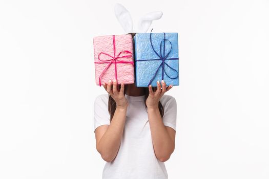 Celebration, holidays and presents concept. Cute Easter bunny female in rabbit ears, hiding her face behind pastel wrapped gift boxes, congratulating, prepare surprise, white background.