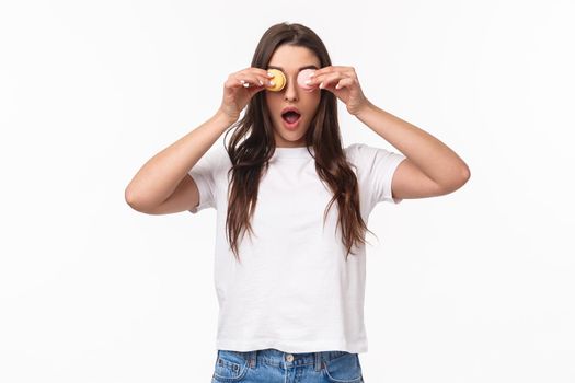 Portrait of amused, excited young cute girl in white t-shirt, holding tasty two macarons over eyes, playing with her food, open mouth and gasping as if surprised, studio background.