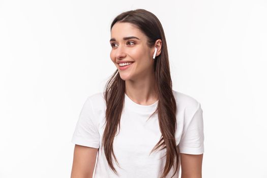 Close-up portrait of lovely, pretty young woman in t-shirt, listening music in wireless headphones, look away with pleased smile, enjoying favorite podcast or songs playlist, white background.