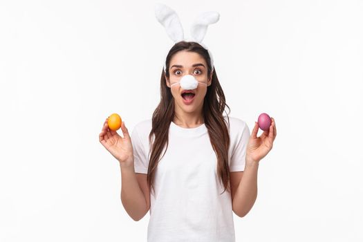 Easter party begins. Portrait of funny and cute, silly young caucasian woman celebrating divine day, wear rabbit ears and nose, holding colored eggs, gasping amused, white background.