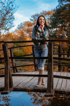 Young happy woman sitanding on a wooden bridge over the little lake in the park and enjoying in a beautiful autumn golden day.