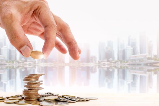 Human hand placing a coin on a pile of coins with blurred skyscraper background, use for business and financial growth concepts.
