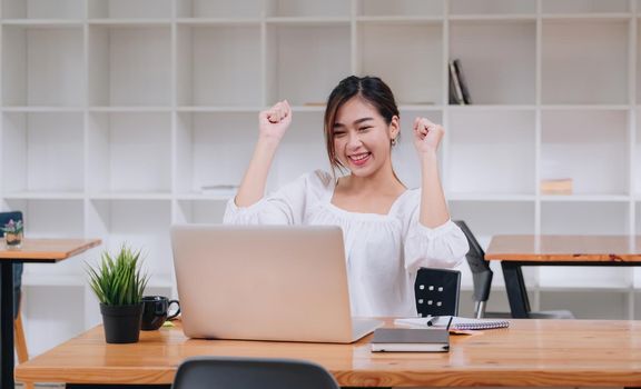 Happy excited asian woman at home workstation triumphing with raised hands, female using laptop computer with online meeting.