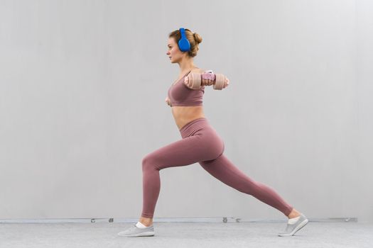 Girl in a tight pink outfit raising dumbbells up in a warrior position. Girl losing doing sports exercises lifting weight listen music in blue wireless headphones. Healthy lifestyle, sport concept.