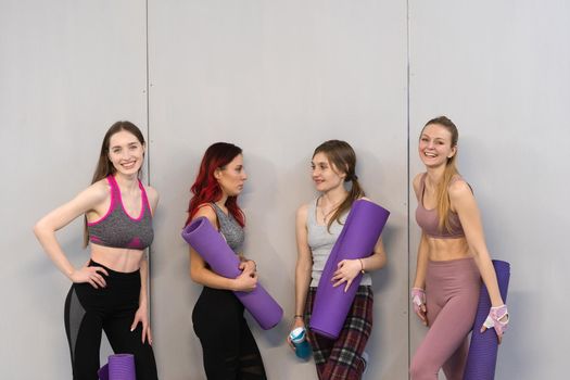 Athletic girls in sports out fits standing next to the wall holding a yoga mat, smiling on camera. Practicing in fitness or yoga studio, working out indoor. Isolated on grey background.