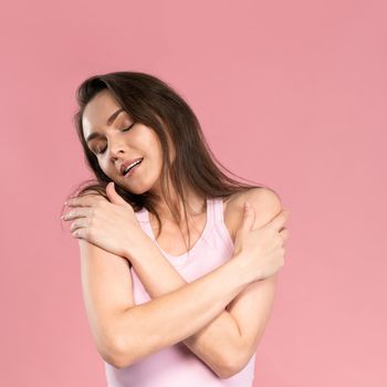 Tender beautiful young woman with no makeup standing with hands crossed on her chest, attractive brunette girl on pink background. Beauty and health care concept.