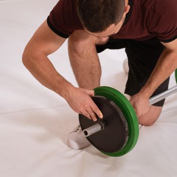 Adding weight to a barbell young man bow on a knee changing black and green plates, equipment for weight training concept. Sports equipment for training. square cropped.