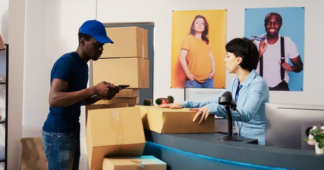 African american deliveryman asking worker to sign distribution report on tablet computer, discussing customer shipping detalis in shopping mall. Employee preparing packages in modern boutique
