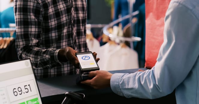 African american man paying for casual wear using mobile phone nfc on pos terminal, buying stylish clothes and accessories in clothing store. Customer shopping for trendy merchandise