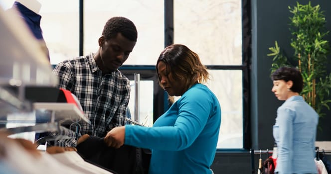 Stylish couple looking at trendy clothes, checking shirt fabric in modern boutique. African american customers shopping for new fashion collection, buying elegant merchandise and accessories