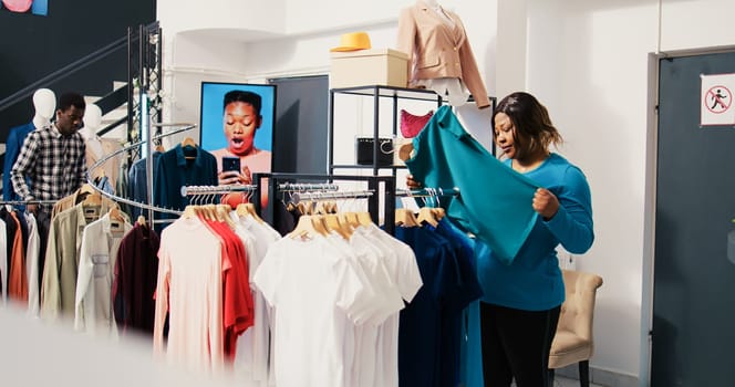 Asian employee helping woman to buy stylish clohtes, discussing shirt material in clothing store. African american customer shopping for fashionable merchandise, looking at new fashion collection