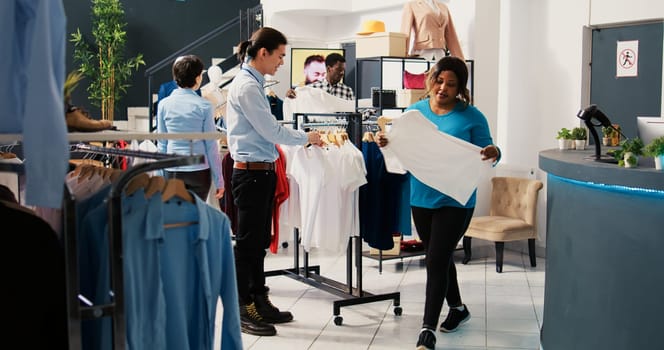 Customer needed help with trendy shirts, discussing merchandise material with asian employee in modern boutique. African american woman shopping for stylish clothes in clothing store