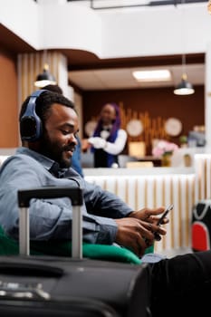 Happy relaxed African American man sitting in hotel lobby listening to music on digital tablet, having video call while waiting for check-in. Tourist wearing headphones looking at device screen