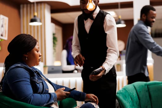 African American man hotel waiter holding POS terminal receiving cashless payment from female guest in lobby. Woman paying for room with phone at reception area. NFC technology in hospitality concept