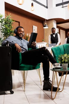 Happy young African American guy using digital tablet and wireless headphones while sitting in hotel lounge area, looking at webcam smiling. Tourist talking by video with family while travelling