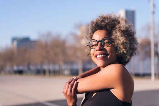 smiling african american woman stretching arms looking at camera in an urban park, concept of health and sportive lifestyle, copy space for text