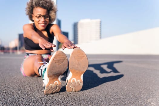 african american woman stretching the legs sitting in an urban park, concept of health and sportive lifestyle, selective focus on the sports shoes and copy space for text