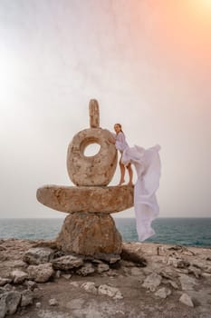 Woman sea stone. A woman stands on a stone sculpture made of large stones. She is dressed in a white long dress, against the backdrop of the sea and sky. The dress develops in the wind