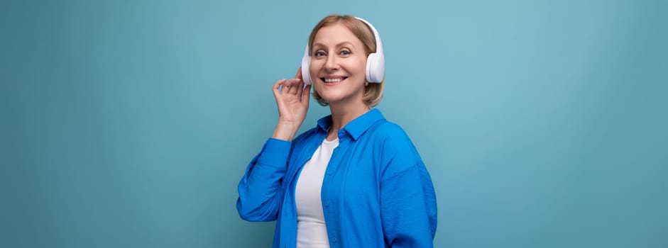 middle age business concept. mature woman listening to audiobook with headphones on blue background copy space.