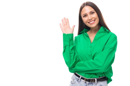 confident smiling young European brown-eyed female model with well-groomed black hair and make-up dressed in a green shirt waves her hand against the background with copy space for advertising.