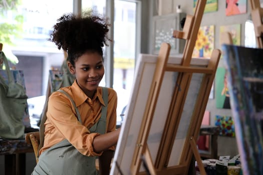 Creative young African American woman wearing apron painting picture with brush on canvas in art studio.