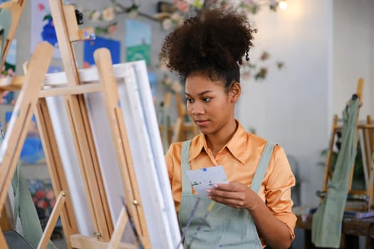 Happy young woman with Afro haired wearing apron painting picture with brush on canvas in art studio.