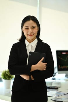 Attractive female office manager or business assistant holding digital tablet standing in modern office.